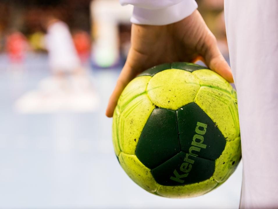 Nahaufnahme einer Hand, die einen grünen Handball mit gelben Elementen hält, im Hintergrund Spieler auf dem Spielfeld.